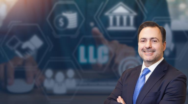 Asher Rubinstein in a dark suit and blue tie, standing with his arms crossed and smiling. The background features a blue-toned business and legal-themed graphic with icons representing finance, partnerships, corporate structures, and institutions, including a prominent “LLC” symbol at the center. The image conveys themes of asset protection, business planning, and corporate law.
