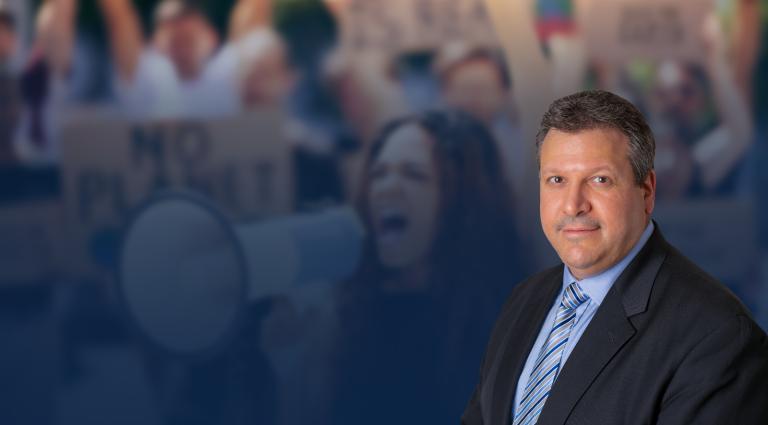 Headshot of Joe Farca in a suit over a blurred dark blue background with an image of a protest.