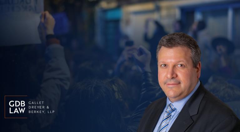 Headshot of Joe Farca in a suit over a blurred dark blue background with an image of a protest.